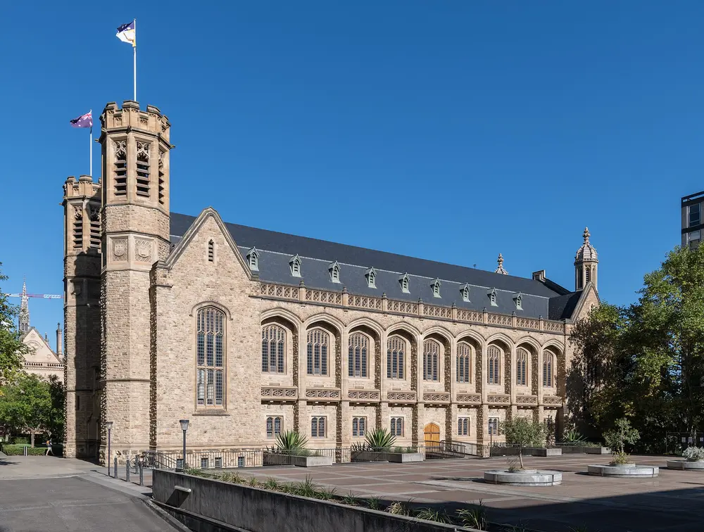 Bonython Hall, the flagship cream-sandstone Gothic great hall of the University of Adelaide (host university of the Adelaide dental school), North Terrace campus — side/east view with crenellated tower and flags under a clear blue sky.