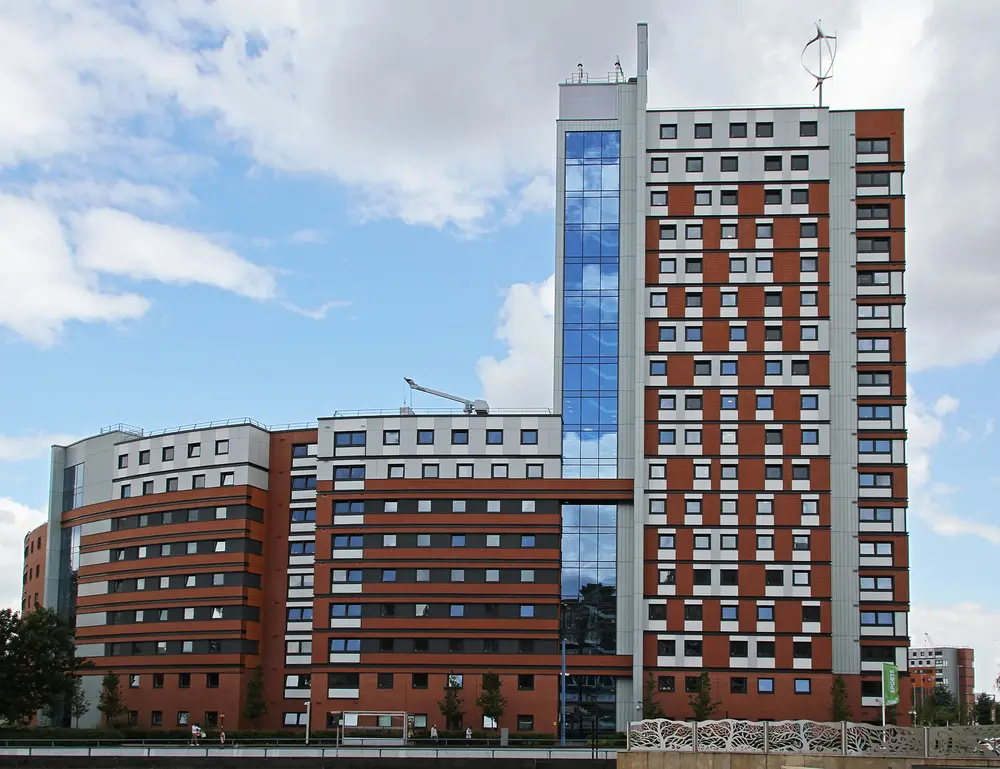 Aston University campus residence towers (red/white/grey clad high-rise with rooftop wind turbine) against a blue sky, Birmingham.
