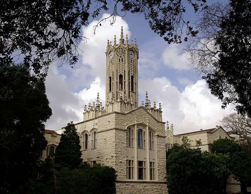 The University of Auckland's iconic Old Arts Building (Clock Tower), a Gothic Revival cream-stone tower, viewed from below against a bright blue sky with dramatic white clouds.