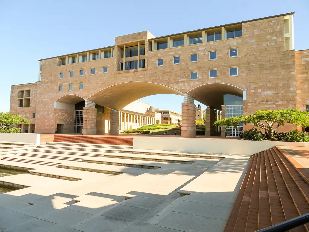 The Arch Building, the iconic sandstone landmark at Bond University's Gold Coast (Robina) campus — daytime frontal exterior, sandstone facade under clear blue sky, empty plaza foreground. Bond University hosts the Bond University Medical Program.