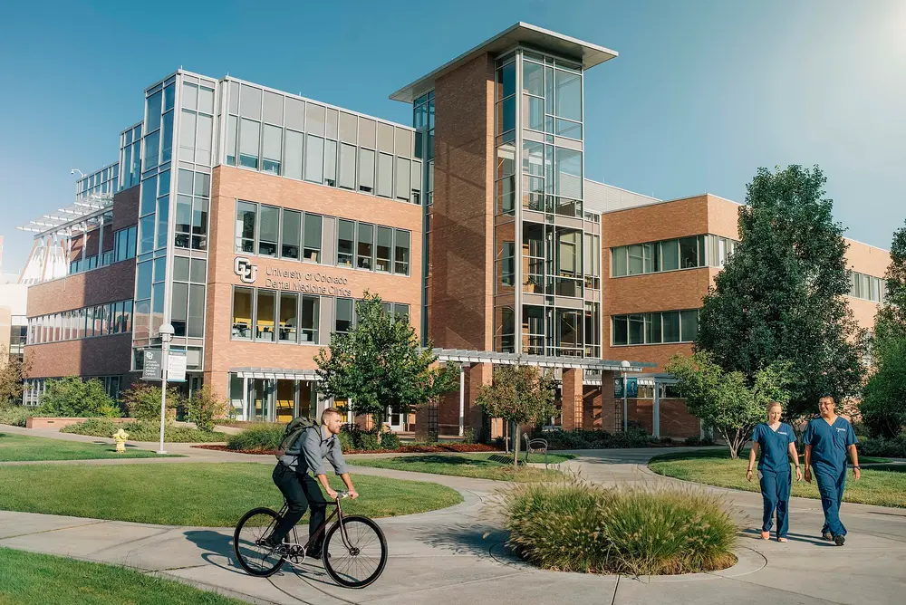 Daytime exterior of the University of Colorado School of Dental Medicine building on the Anschutz Medical Campus, Aurora, CO — brick-and-glass facade with CU/Dental Medicine Clinics signage, blue sky and green lawn.
