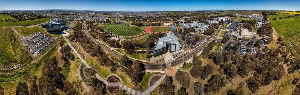 Daytime aerial panorama of Deakin University's Waurn Ponds campus in Geelong, Australia — the campus that hosts the School of Medicine — showing the modern angular academic building, sports oval and blue sky.