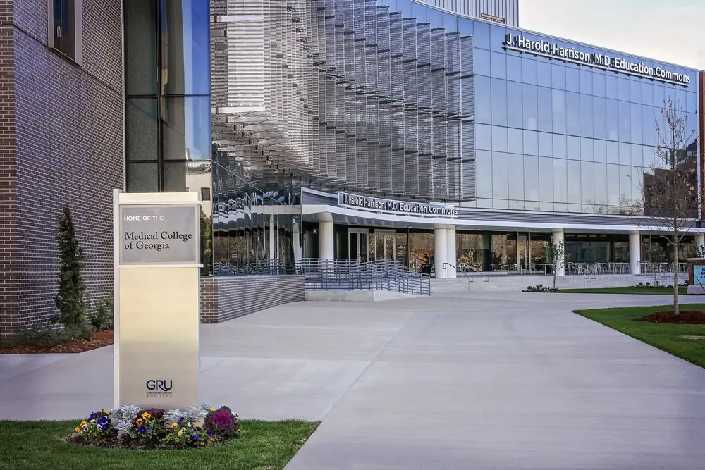 Daytime exterior of the J. Harold Harrison, M.D. Education Commons, the modern glass academic building on Augusta University's Health Sciences campus in Augusta, GA — home to the Medical College of Georgia and the Dental College of Georgia.