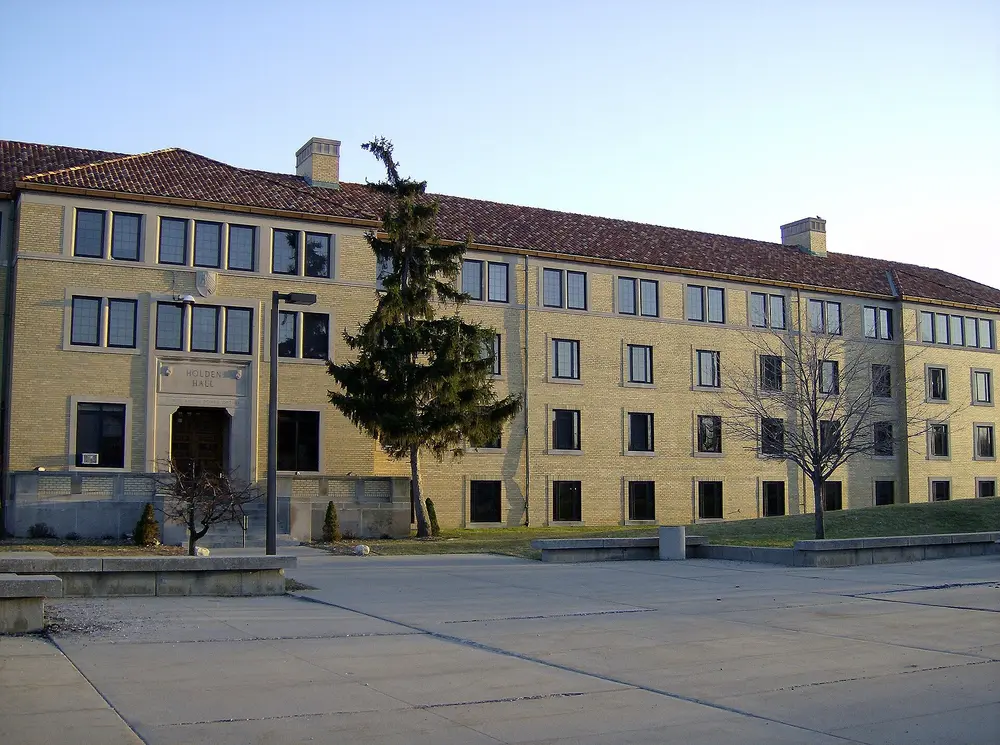 Holden Hall on the University of Detroit Mercy McNichols campus, Detroit, Michigan — a symmetric collegiate stone/brick building with red tile roof under a clear blue sky, wide daytime exterior with an open plaza foreground.