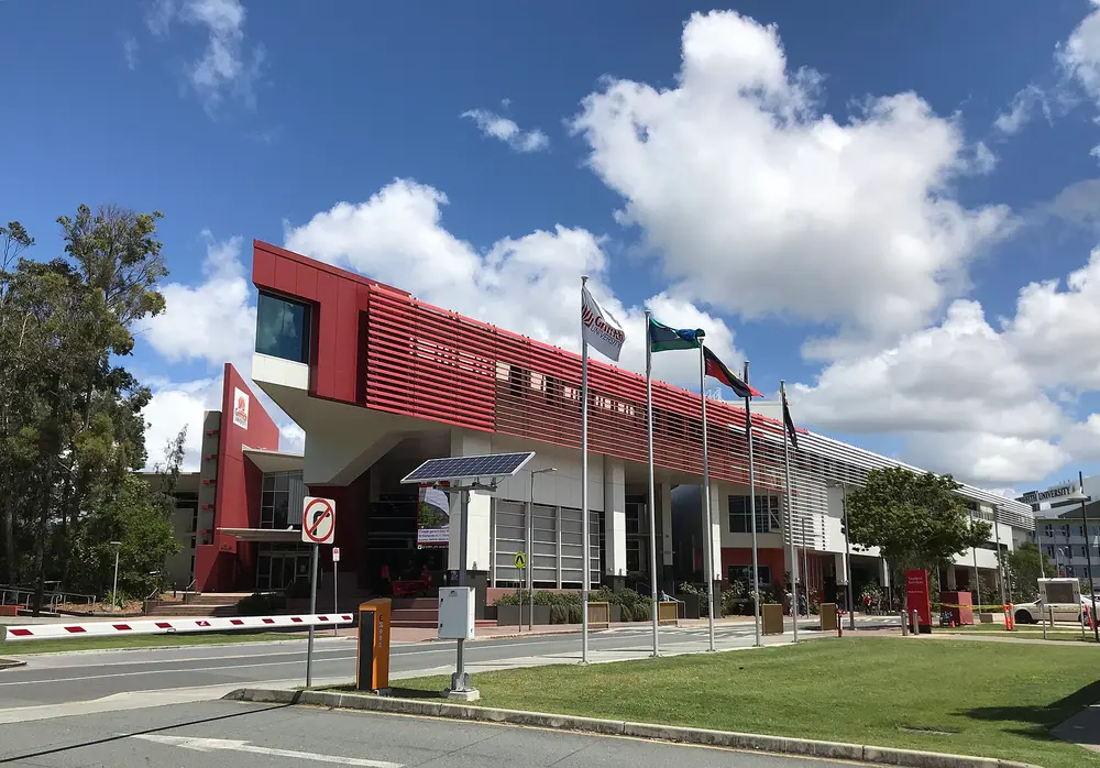 Exterior of the iconic red-and-white Griffith University Gold Coast Campus building (Library/G-block), the campus that hosts Griffith's School of Medicine and Dentistry, under a bright blue sky with Griffith University branding and flags.