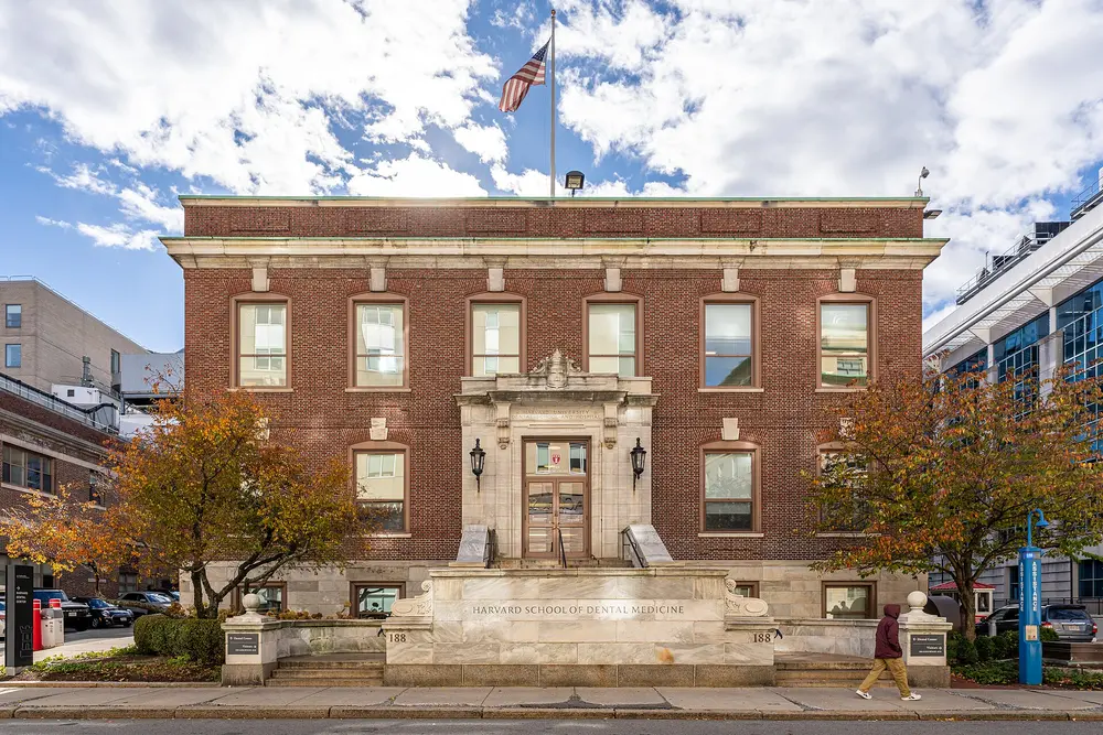 Symmetric front exterior of the Harvard School of Dental Medicine building at 188 Longwood Avenue, Boston, MA, with the school name carved in stone, blue sky and autumn trees.