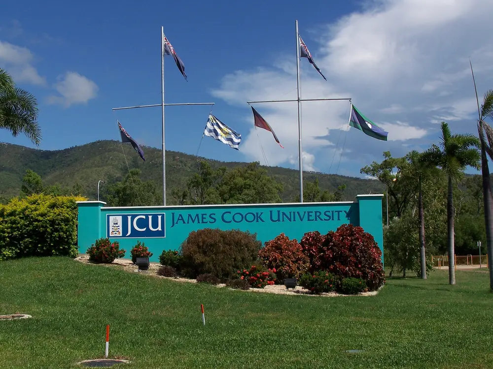 The James Cook University main entrance monument at the Townsville (Douglas) campus, with the JCU sign and multiple flags, Mount Stuart bushland behind, and a clear blue daytime sky.