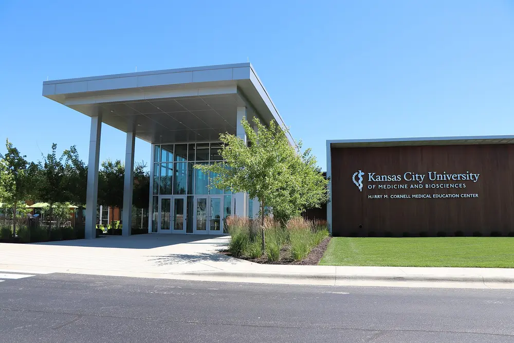 Exterior of the Kansas City University Joplin campus (Harry M. Cornell Medical Education Center) — the campus housing KCU's College of Dental Medicine — a modern glass-and-wood building under a clear blue sky.