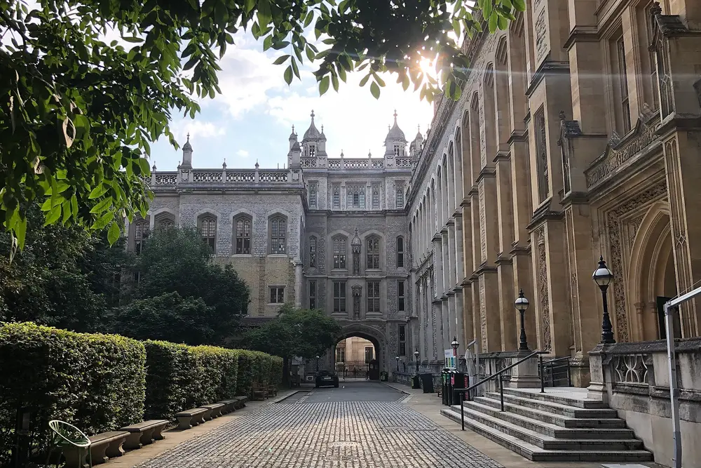 Daytime exterior view of King's College London's neo-Gothic Strand campus quad, with the pinnacled stone building and an ornate Gothic archway along a cobbled courtyard lane.