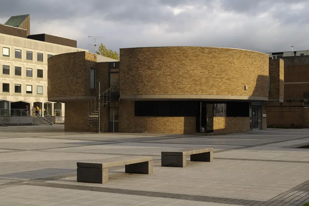 A campus square at Lancaster University (Bailrigg) — a round yellow-brick building lit by warm evening sun under a dramatic sky, with a clean paved plaza and benches in the foreground.