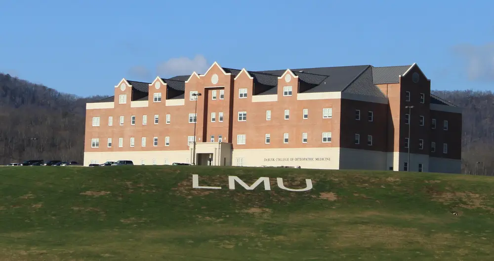 DeBusk College of Osteopathic Medicine, the flagship health-sciences building on Lincoln Memorial University's main campus — red-brick collegiate building with blue sky, green lawn spelling "LMU", framed by mountains.