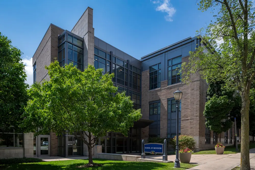 Daytime exterior of the Marquette University School of Dentistry building in Milwaukee, WI, with the "School of Dentistry" sign, framed by green trees under a clear blue sky.