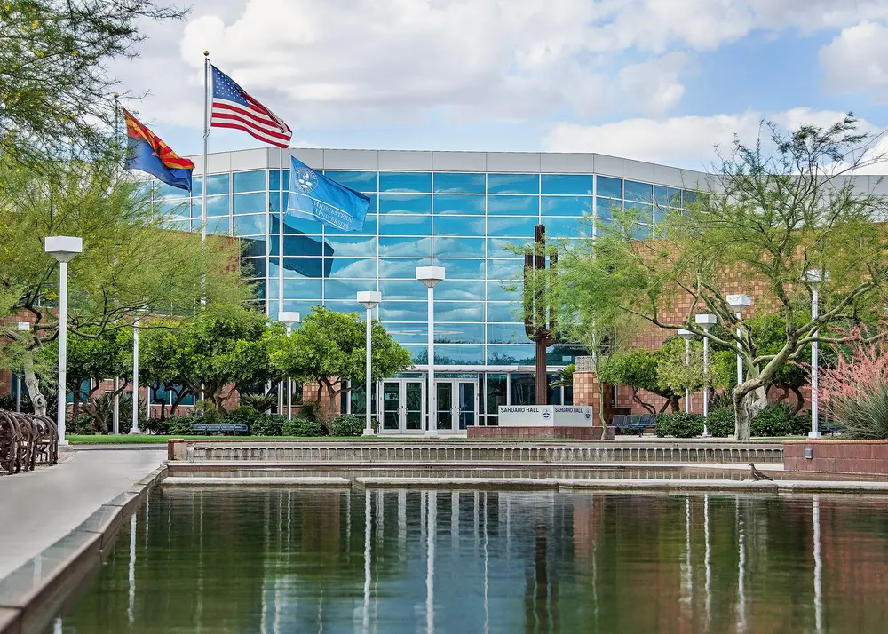 Sahuaro Hall, the flagship blue-glass academic building at Midwestern University's Glendale, Arizona campus (home of the College of Dental Medicine-Arizona), seen straight-on across the reflecting pool with US, Arizona, and Midwestern University flags.