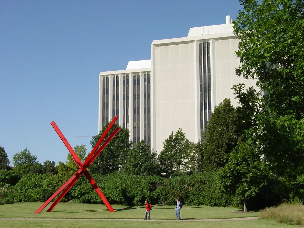 Daytime exterior of the University of Nebraska-Lincoln campus: the tall cream Hamilton Hall science tower rising behind the Sheldon sculpture garden, deep blue sky, green trees and two students walking.