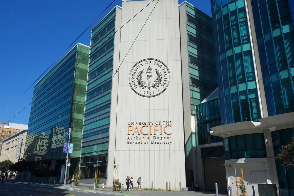 Daytime street-level exterior of the University of the Pacific Arthur A. Dugoni School of Dentistry building in San Francisco, CA — modern glass facade with the University of the Pacific seal and school name, under a clear blue sky.