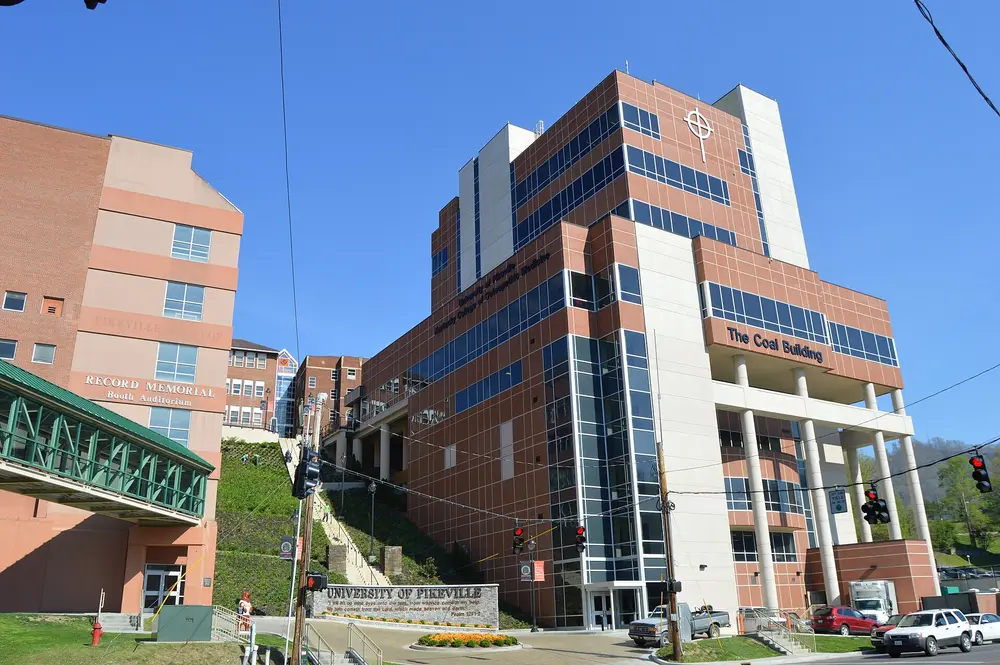 Daytime exterior of the University of Pikeville campus, showing the health-sciences buildings including "The Coal Building" (Kentucky College of Osteopathic Medicine), Record Memorial / Booth Auditorium, and the University of Pikeville entrance sign, under a clear blue sky.