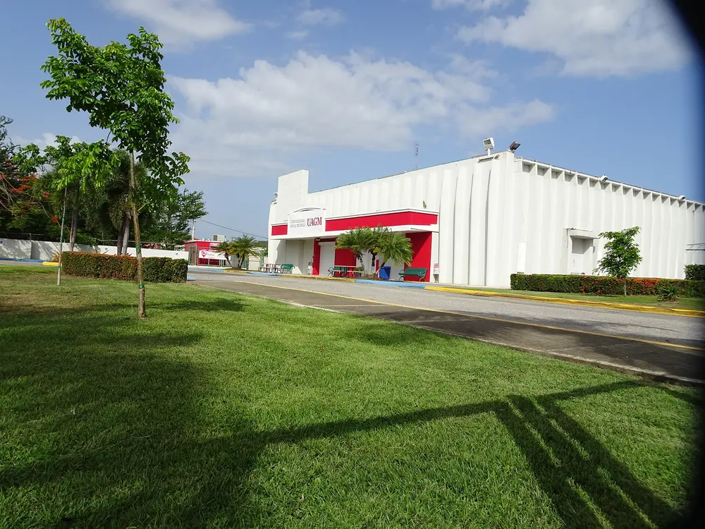 Daytime exterior of a Universidad Ana G. Méndez (UAGM) campus building — white facade with red accents, university name/branding on the entrance, blue sky and green lawn.
