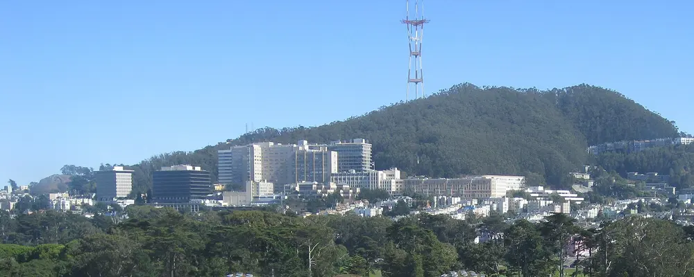 Daytime landscape view of the UCSF Parnassus Heights campus / medical center buildings on the hillside below Mount Sutro and Sutro Tower in San Francisco, CA — the campus that hosts the UCSF School of Dentistry.