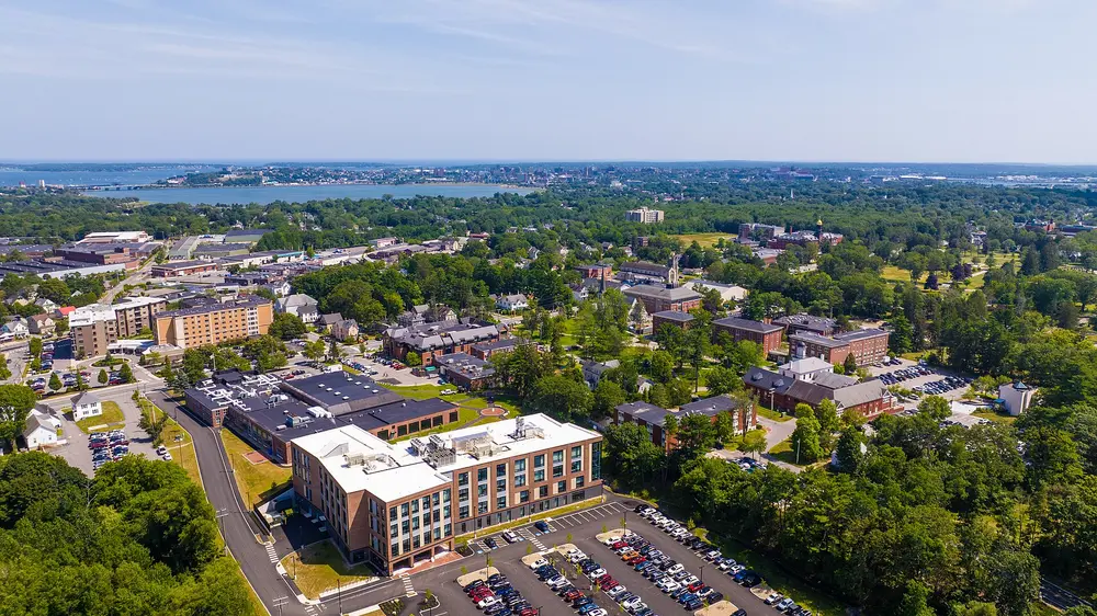 Summer daytime aerial of the University of New England's Portland Campus for the Health Sciences (Portland, ME) — the campus that houses the College of Dental Medicine — with the campus quad, Back Cove and Portland skyline behind under a clear blue sky.