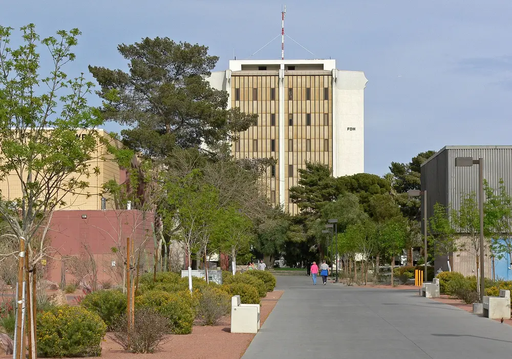 Ground-level daytime view of the University of Nevada, Las Vegas (UNLV) main campus, showing a landmark campus tower (FDH) rising above a landscaped pedestrian walkway under a clear blue sky.