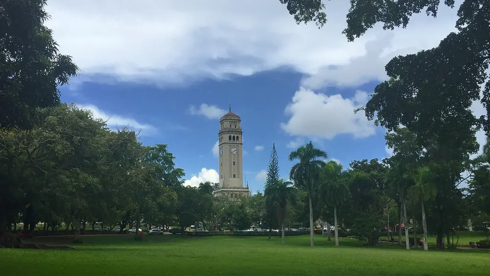 The iconic Roosevelt Tower (Torre) clock tower of the University of Puerto Rico, Río Piedras Campus, viewed across a green lawn under a dramatic blue sky, in San Juan, PR.