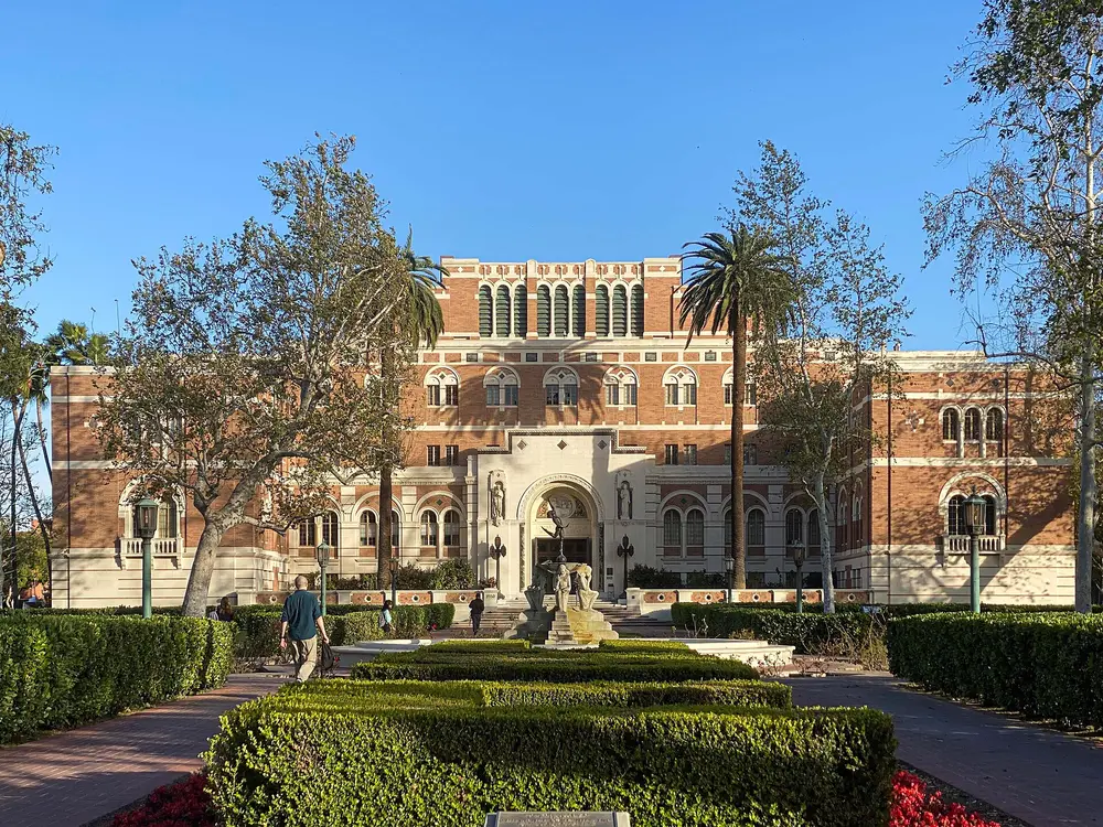 The iconic Edward L. Doheny Jr. Memorial Library on the main campus of the University of Southern California (USC) in Los Angeles — the parent university of the Herman Ostrow School of Dentistry — shot head-on in daytime with a bright blue sky, framed by trees, palms and manicured hedges.
