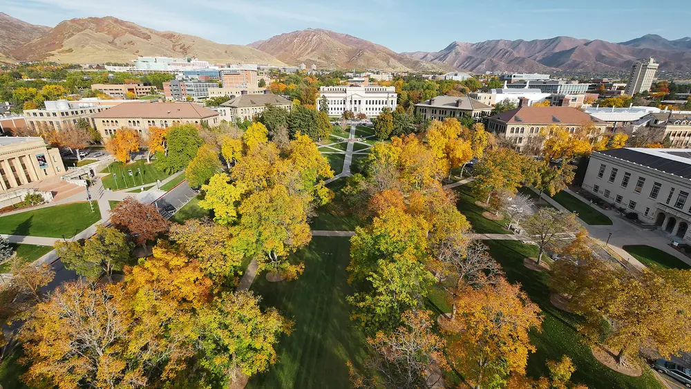 Aerial daytime view of the University of Utah's historic Presidents Circle in Salt Lake City — the neoclassical Park Building framed by an autumn tree-lined quad with the Wasatch mountains behind.