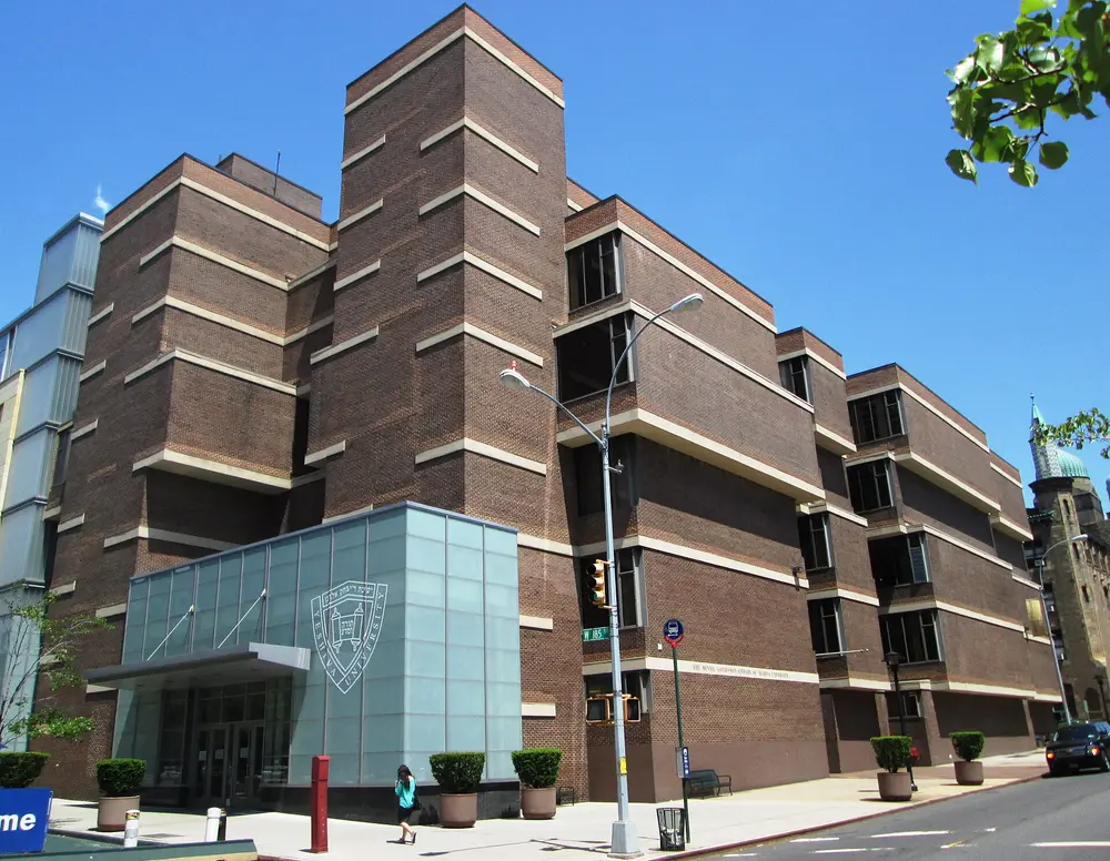 Yeshiva University's Mendel Gottesman Library on the Wilf Campus (Washington Heights, Manhattan, NYC) — the parent university of the College of Dental Medicine — under a bright blue daytime sky, YU seal on the glass entrance and Zysman Hall dome visible in the background.